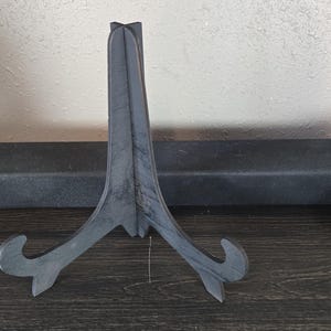 May include: A black metal easel stand with a simple design. The stand is made of a single piece of metal and has a triangular shape.