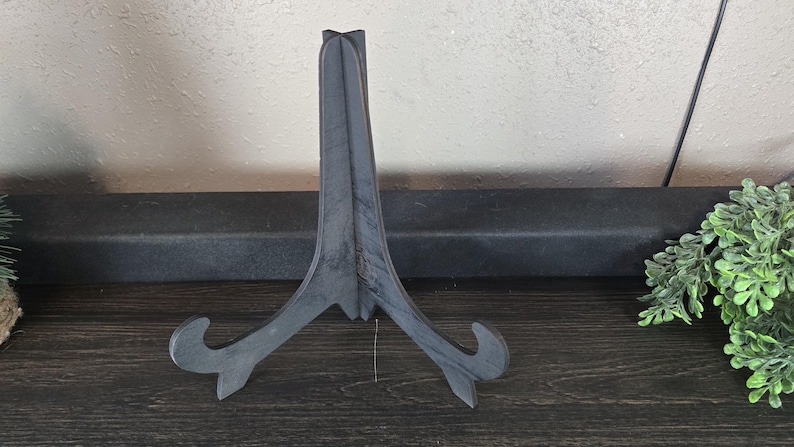 May include: A black metal easel stand with a simple design. The stand is made of a single piece of metal and has a triangular shape.