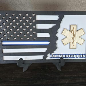 May include: A black framed wooden American flag with a blue stripe and a white background. The flag is partially cut out to reveal a wooden medical symbol with a snake and a staff. The name "McNaughton" is written below the symbol.