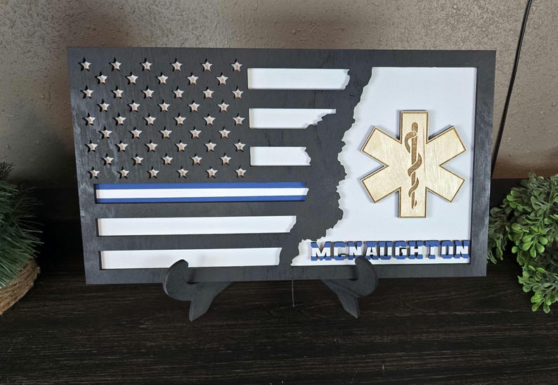 May include: A black framed wooden American flag with a blue stripe and a white background. The flag is partially cut out to reveal a wooden medical symbol with a snake and a staff. The name "McNaughton" is written below the symbol.