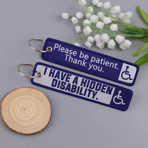 May include: Two embroidered keyrings. One is navy blue with white text that reads "Please be patient. Thank you." and a wheelchair symbol. The other is white with navy text that reads "I HAVE A HIDDEN DISABILITY." and a wheelchair symbol.