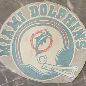 May include: A white and blue Miami Dolphins logo with a dolphin jumping out of a helmet. The logo is surrounded by a circle with the words "Miami Dolphins" in blue.