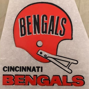 May include: A vintage iron-on patch featuring the Cincinnati Bengals logo. The logo is a red football helmet with a black outline and the word "BENGALS" in black lettering. The word "CINCINNATI" and the word "BENGALS" in red lettering are below the helmet.
