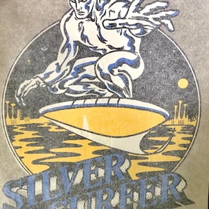 May include: A vintage Silver Surfer t-shirt featuring a graphic of the Silver Surfer riding his surfboard in the night sky. The text "SILVER SURFER" is printed in blue and white letters below the graphic. The copyright information "© 1978 MARVEL COMICS GROUP ALL RIGHTS RESERVED" is printed in small letters at the bottom of the shirt.