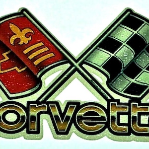 May include: A metallic decal featuring the Chevrolet Corvette logo with two crossed flags. One flag is red with a gold fleur-de-lis and horizontal stripes, while the other is a black and white checkered flag. The word corvette is written below.