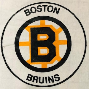 May include: A white circle with a black outline and the words "BOSTON" and "BRUINS" in black text. Inside the circle is a black "B" with an orange outline, surrounded by a yellow and black hockey puck design.