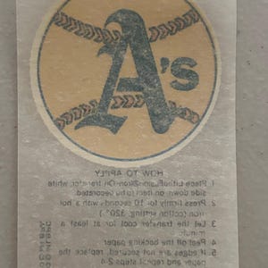 May include: A vintage iron-on transfer featuring the Oakland Athletics logo. The logo is a large, stylized "A" with "'s" in a baseball-themed design, set against a tan background. Instructions are printed below.