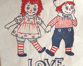 Puede incluir: Dos muñecas de trapo, una con un vestido de cuadros rojos y blancos y la otra con una camisa y pantalones de cuadros azules y blancos, se toman de la mano y sonríen. La palabra "LOVE" está impresa debajo en azul.