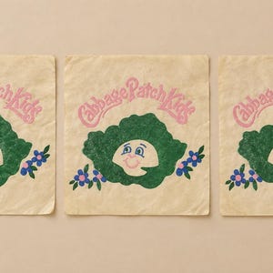 May include: Three square beige fabric patches featuring the pink text Cabbage Patch Kids above a green cabbage leaf graphic. The cabbage leaf contains a smiling face with blue eyes and pink cheeks, flanked by small blue flowers with green leaves.