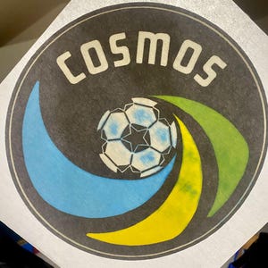 May include: A black and white circular logo with the word "COSMOS" in white lettering. A white soccer ball with a blue star in the center is surrounded by a blue and yellow crescent shape and a green arc.