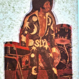 May include: A vintage image of a musician in a black jumpsuit adorned with gold stars, moons, and circles. The word "SLY" is emblazoned across the waist. The musician is smiling, with a microphone and a keyboard in the foreground, and a drum set in the background.