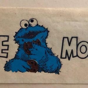 May include: A blue Cookie Monster character with big eyes eating a cookie. The text "COOKIE MONSTER" is written in black letters on a white background.