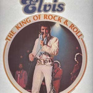 May include: A vintage image featuring Elvis Presley on stage, dressed in a white jumpsuit with intricate embellishments. The image is framed by a circular design with the text "The King of Rock & Roll" and "1935-1977". The word "Elvis" is displayed at the top.