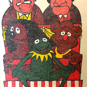 May include: A colorful illustration featuring a green frog character with a yellow collar sitting in front of a red theater balcony. Behind the frog are three brown, furry puppet characters and two older men with red faces, all positioned within a red structure.