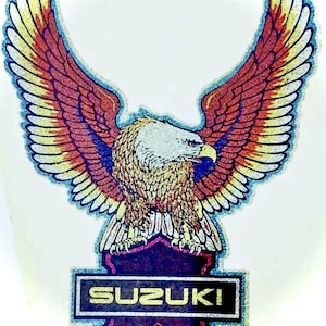 May include: A glittery decal featuring a bald eagle with outstretched wings in shades of red, yellow, and blue. Below the eagle is a dark purple shield-shaped emblem containing the word SUZUKI in yellow text and a small logo.