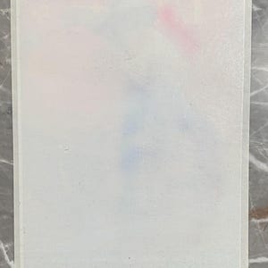 May include: A rectangular, white paper with a faint watercolour design in pink, blue, and purple hues. The paper has a thin, white border and is set against a grey marble surface.