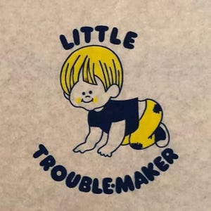 May include: A beige tote bag with a blue and yellow cartoon illustration of a baby with the text "LITTLE TROUBLEMAKER" printed on the front.