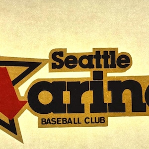 May include: A vintage Seattle Mariners baseball team logo. The logo features a gold star with a black outline and a red "M" shape inside. The words "Seattle Mariners Baseball Club" are in black and gold.