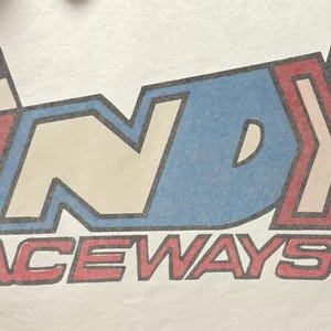 May include: A white t-shirt with a vintage-style logo featuring the word "INDY" in blue, red, and white with a checkered flag design. The text "RACEWAYS" is below the logo in red and black.