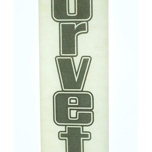 May include: A vertical decal featuring the word CORVETTE in bold, dark gray block letters with a white outline. Above the text are two crossed flags, one with a checkered pattern and the other with a stylized emblem, all set on a light background.