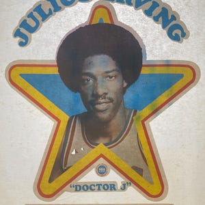 May include: A vintage basketball t-shirt featuring a photo of Julius Erving, nicknamed "Doctor J", wearing a Philadelphia 76ers jersey. The shirt has a large yellow star with a blue center and red outline. The text "Julius Erving" is in a blue arc above the star, and "Philadelphia" is in a blue arc below the star. The text "Doctor J" is in a white arc below the star.