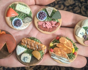 Puede incluir: Tablas de embutidos en miniatura con quesos, carnes, aceitunas y pan. Cada tabla redonda de madera tiene alimentos detallados. Las tablas se sostienen en una mano, mostrando su pequeña escala. Aproximadamente 3 cm de diámetro.