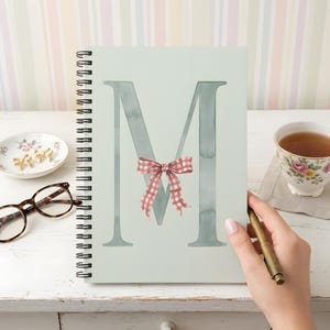 May include: A spiral-bound notebook with a light sage green cover featuring a large, dark sage letter M in the center. A small, pink and white gingham patterned bow is illustrated over the middle of the letter M. The notebook is rectangular.