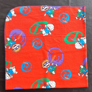 Può includere: Coperta per neonati in tessuto rosso con un motivo ripetitivo di personaggi dei cartoni animati e la parola "Chuckie" in un cerchio viola.