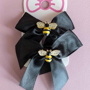 May include: Two black satin hair bows with gold and rhinestone bee embellishments. The bows are attached to a white and pink bow-shaped card. The bees have yellow and black stripes.