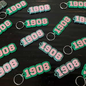 May include: Keychains with the number "1908" in pink and white, outlined in green, attached to silver keyrings. The keychains are scattered on a dark surface.