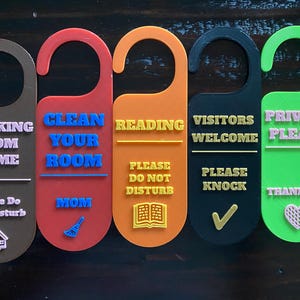 Può includere: Una collezione di cinque cartelli per porta colorati con vari messaggi. I messaggi includono "Working From Home", "Clean Your Room", "Reading", "Visitors Welcome" e "Privacy Please". Ogni cartello ha un colore e un design unici.