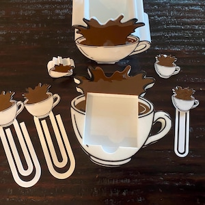 May include: A collection of coffee-themed paperclips, bookmarks, and a card holder. The designs feature white coffee cups with brown coffee spills. The items are arranged on a dark wooden surface.