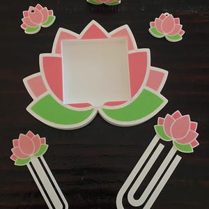 May include: A collection of lotus flower-themed accessories in pink, green, and white. Includes a large lotus-shaped frame, two paperclip bookmarks, two small charms, and a 3D lotus flower. The items are arranged on a dark wooden surface.
