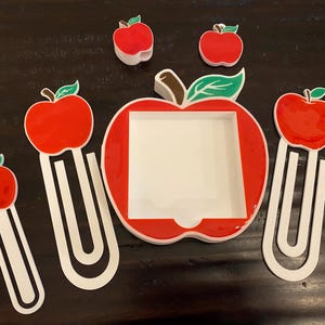 May include: A collection of red apple-shaped paper clips and a large apple-shaped holder. The apples have green leaves and brown stems. The paper clips are white with a red apple on top. The holder has a white interior.