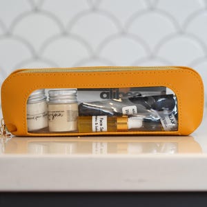 May include: A bright orange cosmetic bag with a clear window, filled with small jars and bottles. The bag contains skincare products, including face serum and face cream. The bag has a gold zipper and a gold charm.