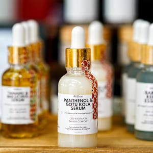 May include: A clear glass serum bottle with a white label featuring the text "AliBee PANTHENOL & GOTU KOLA SERUM". The bottle has a gold cap and a white dropper. The label also includes "DEEP HYDRATION BARRIER COMFORT".
