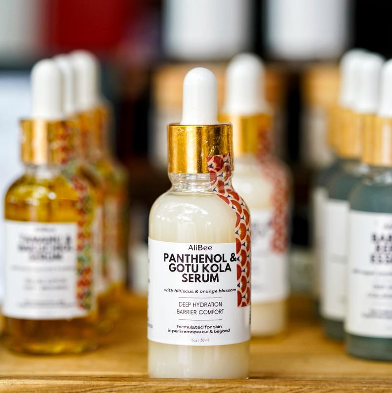 May include: A clear glass serum bottle with a white label featuring the text "AliBee PANTHENOL & GOTU KOLA SERUM". The bottle has a gold cap and a white dropper. The label also includes "DEEP HYDRATION BARRIER COMFORT".