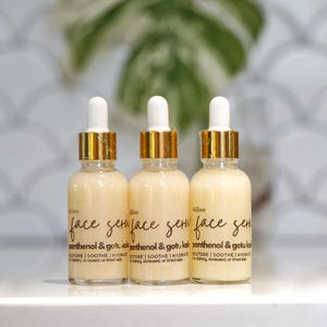 May include: Three glass serum bottles with white droppers and gold-colored collars. The bottles are filled with a light yellow liquid and have black labels with the words "face serum" and "panthenol & gotu kola".