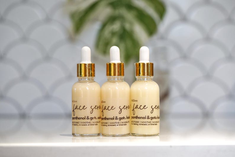 May include: Three glass serum bottles with white droppers and gold-colored collars. The bottles are filled with a light yellow liquid and have black labels with the words "face serum" and "panthenol & gotu kola".