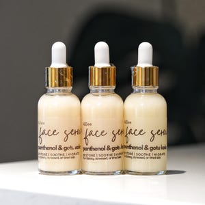 May include: Three glass bottles of face serum with white and gold accents. Each bottle has a white dropper and a label that reads "face serum" and "panthenol & gotu kola". The serum is designed to restore, soothe, and hydrate thirsty, stressed, or tired skin.