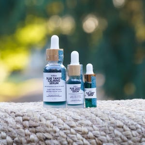 K&ouml;nnte beinhalten: Drei Glasflaschen mit Pipette f&uuml;r Blue Tansy Calming Serum in verschiedenen Gr&ouml;&szlig;en. Jede Flasche hat einen wei&szlig;en Pipettenaufsatz, einen hellen Holzring mit blau-wei&szlig;em Muster und ein Etikett mit dem Text Blue Tansy Calming Serum.