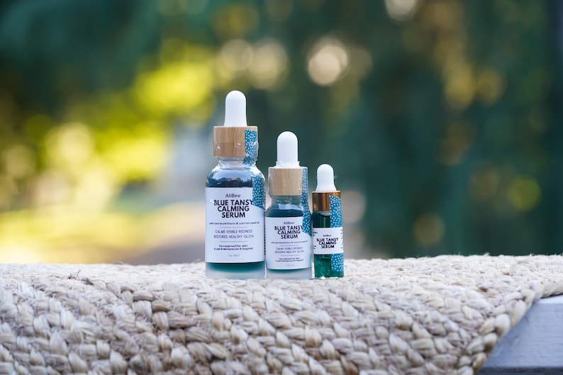 K&ouml;nnte beinhalten: Drei Glasflaschen mit Pipette f&uuml;r Blue Tansy Calming Serum in verschiedenen Gr&ouml;&szlig;en. Jede Flasche hat einen wei&szlig;en Pipettenaufsatz, einen hellen Holzring mit blau-wei&szlig;em Muster und ein Etikett mit dem Text Blue Tansy Calming Serum.