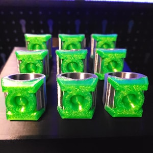 May include: Nine green and silver shot glasses arranged in a grid. Each shot glass has a square, green exterior with a circular design and a silver interior. The shot glasses are displayed on a black surface.