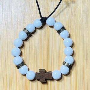 May include: A white beaded bracelet with decorative metal spacers and a wooden cross pendant. The bracelet is strung on a black cord and is displayed against a light brown wooden background.