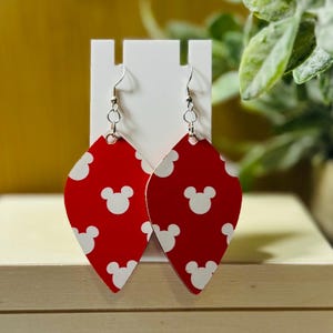 Puede incluir: Pendientes rojos y blancos con tem&aacute;tica de Mickey Mouse. Los pendientes tienen forma de l&aacute;grima con un fondo rojo y patrones de cabeza de Mickey Mouse blancos. Tienen alambres para las orejas de gancho de pez plateados y se exhiben en un soporte blanco.