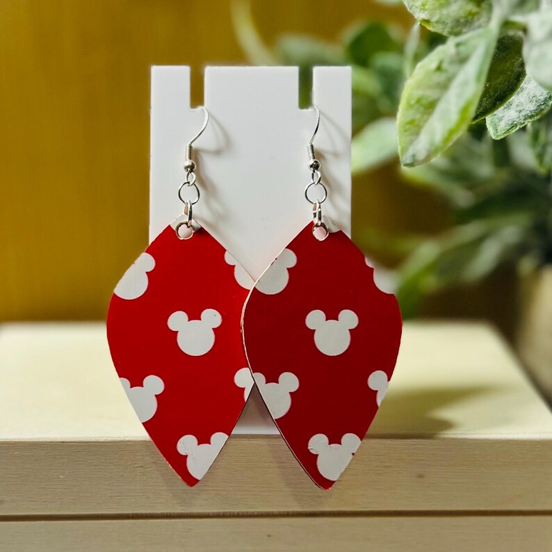 Puede incluir: Pendientes rojos y blancos con tem&aacute;tica de Mickey Mouse. Los pendientes tienen forma de l&aacute;grima con un fondo rojo y patrones de cabeza de Mickey Mouse blancos. Tienen alambres para las orejas de gancho de pez plateados y se exhiben en un soporte blanco.