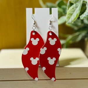 Puede incluir: Pendientes rojos y blancos estampados. Estos pendientes colgantes presentan un fondo rojo con formas de cabeza de Mickey Mouse blancas. Los pendientes se muestran en un soporte blanco. Los ganchos son de color plateado.
