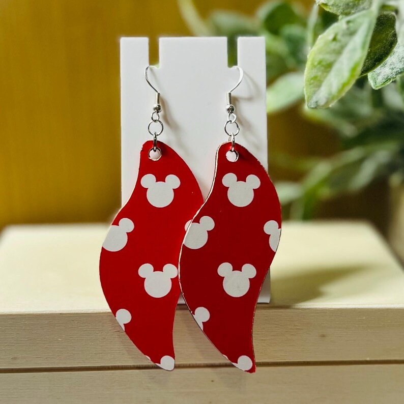 Puede incluir: Pendientes rojos y blancos estampados. Estos pendientes colgantes presentan un fondo rojo con formas de cabeza de Mickey Mouse blancas. Los pendientes se muestran en un soporte blanco. Los ganchos son de color plateado.