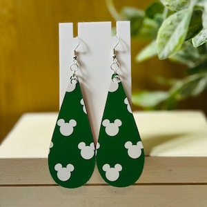 Puede incluir: Un par de pendientes verdes en forma de l&aacute;grima con patrones de cabezas de Mickey Mouse blancas. Los pendientes se exhiben en un soporte blanco, con ganchos plateados. El fondo es verde y una superficie amarilla.