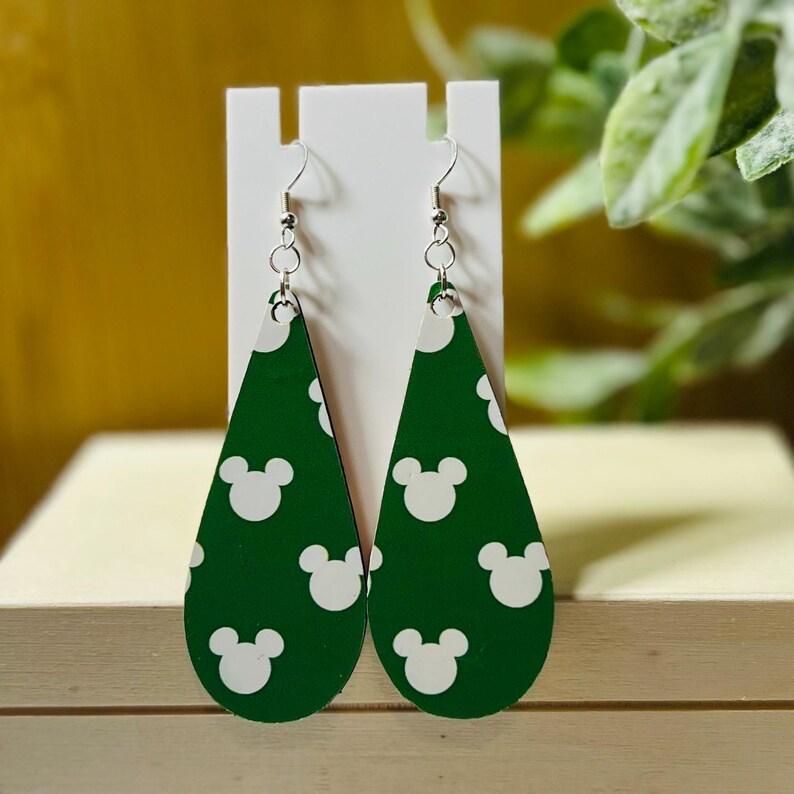 Puede incluir: Un par de pendientes verdes en forma de l&aacute;grima con patrones de cabezas de Mickey Mouse blancas. Los pendientes se exhiben en un soporte blanco, con ganchos plateados. El fondo es verde y una superficie amarilla.